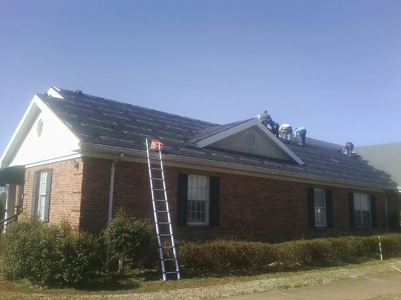 Crew tearing shingles off a brick ranch roof with a ladder set — Emergency Roof Repair work by our crew in Sun Prairie