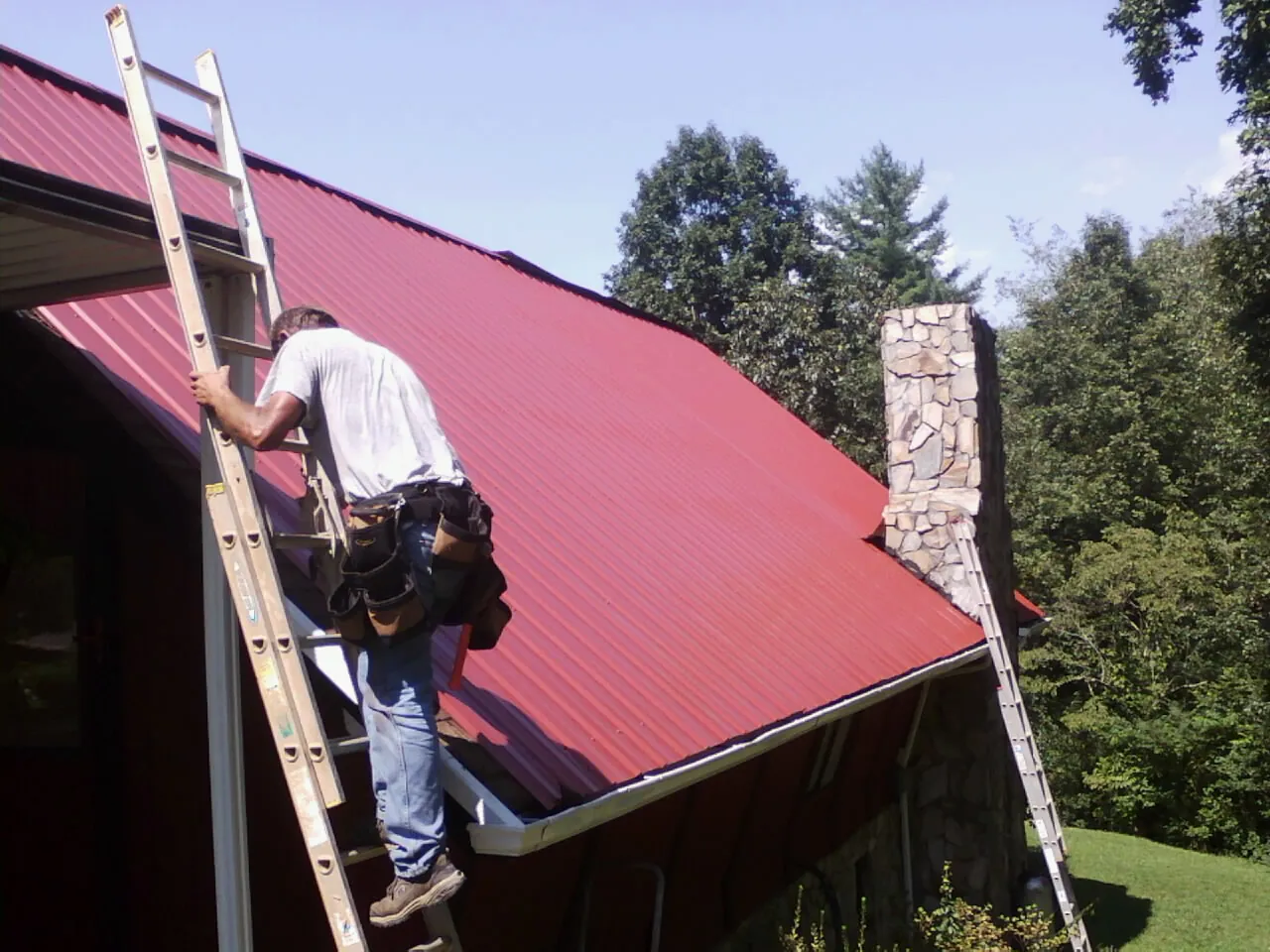 Roofer on a ladder beside a red metal roof and stone chimney — Soffit & Fascia Repair by Roofing Brothers in Sun Prairie